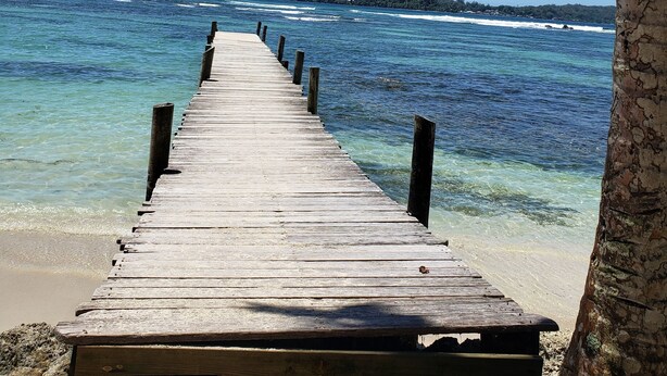 Eine Holzstege, der sich in klares, blaues Wasser erstreckt, mit einem Sandstrand und einer fernen Insel.