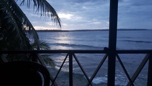 Blick von einer überdachten Terrasse auf den Ozean, mit Geländer und Palmenblättern.
