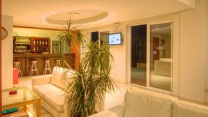 Ein Wohnzimmer mit einer Couch, einem Couchtisch, einer Bar mit Hockern, einer großen Kübelpflanze und einem an der Wand befestigten Fernseher.