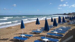 Ein Strand mit Reihen aus blauen Liegestühlen und Sonnenschirmen, einem sandigen Ufer und dem Meer im Hintergrund.