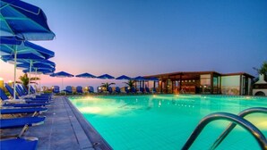 Ein Schwimmbecken mit blauen Liegestühlen und Sonnenschirmen, ein überdachter Sitzbereich und ein Gebäude mit großen Fenstern.