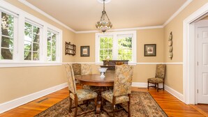 A dining room with a round table, floral chairs, a chandelier, and large windows with a view of trees.