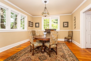 A dining room with a round table, floral chairs, a chandelier, and large windows with a view of trees.
