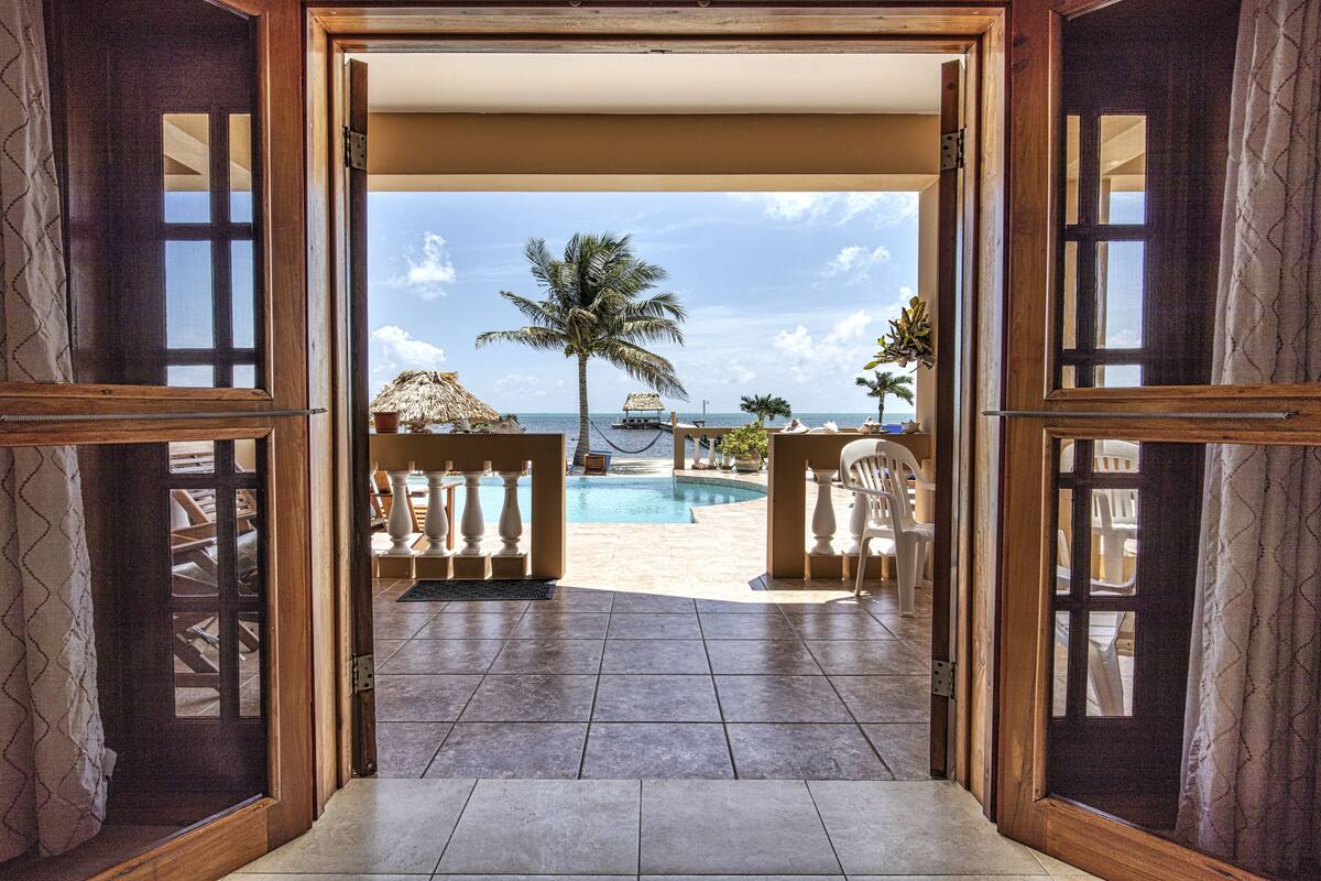 An open doorway leading to a pool area with a view of the beach, palm trees, and a thatched-roof structure.