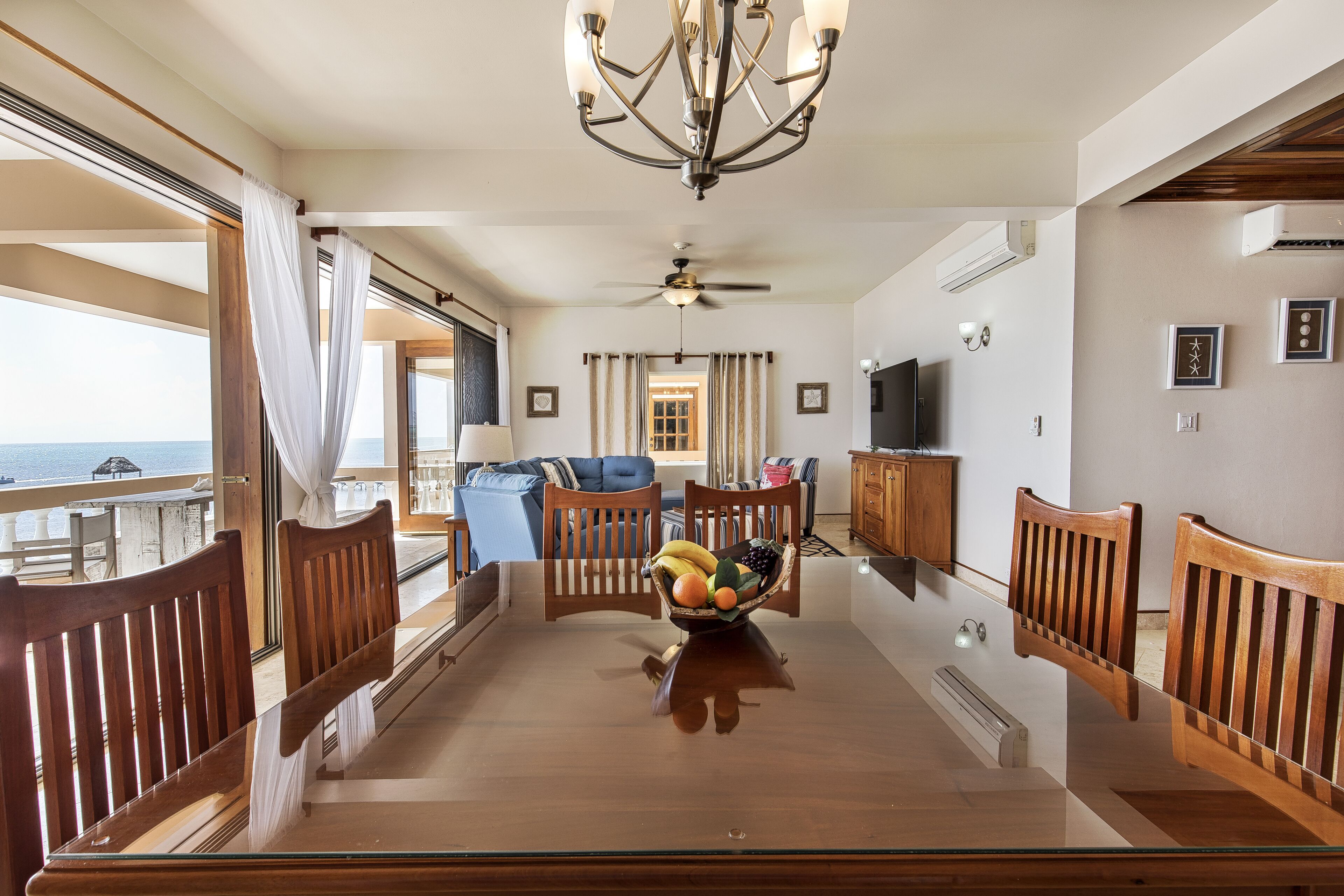 A dining area with a glass table, wooden chairs, a fruit bowl, and a view of the sea.