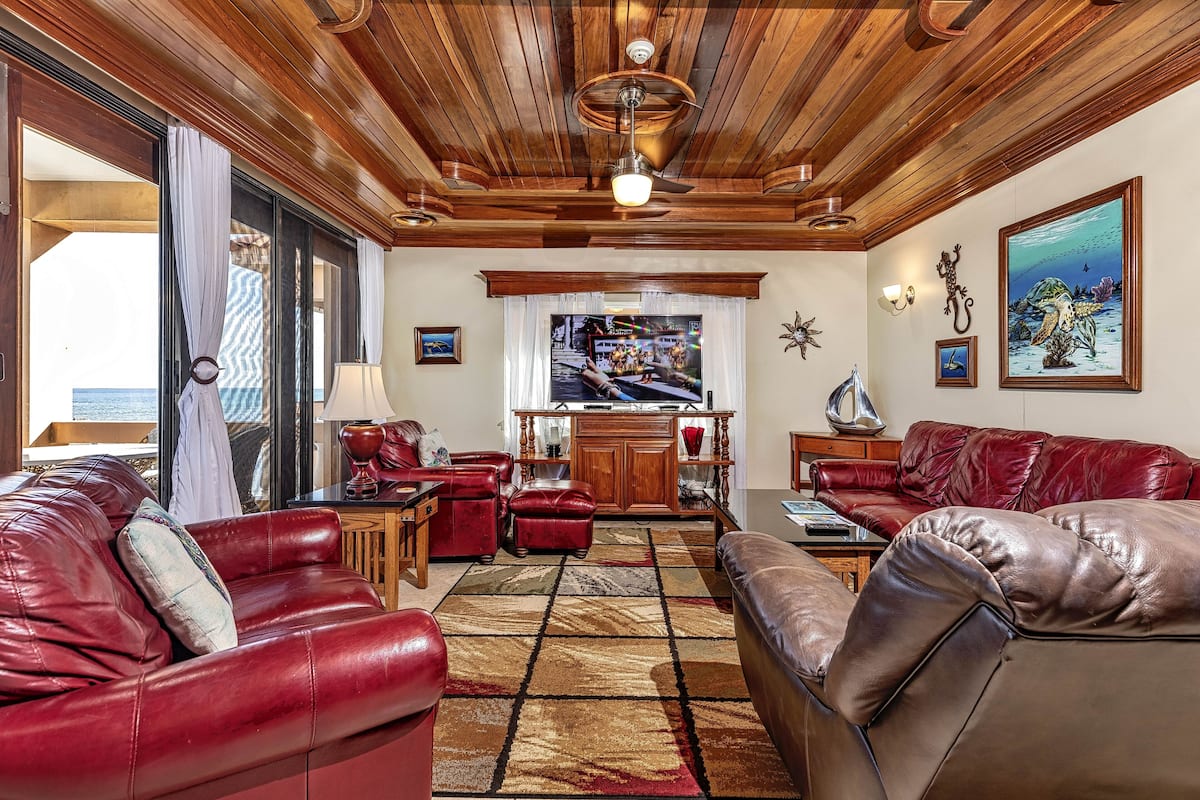 A spacious living room with leather sofas, a wooden ceiling, and a large television.