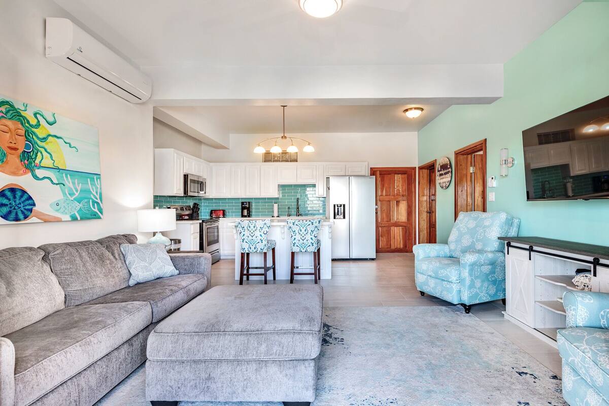 A modern living room with a grey sofa, a blue patterned armchair, and a kitchen area with white cabinets and a teal backsplash.