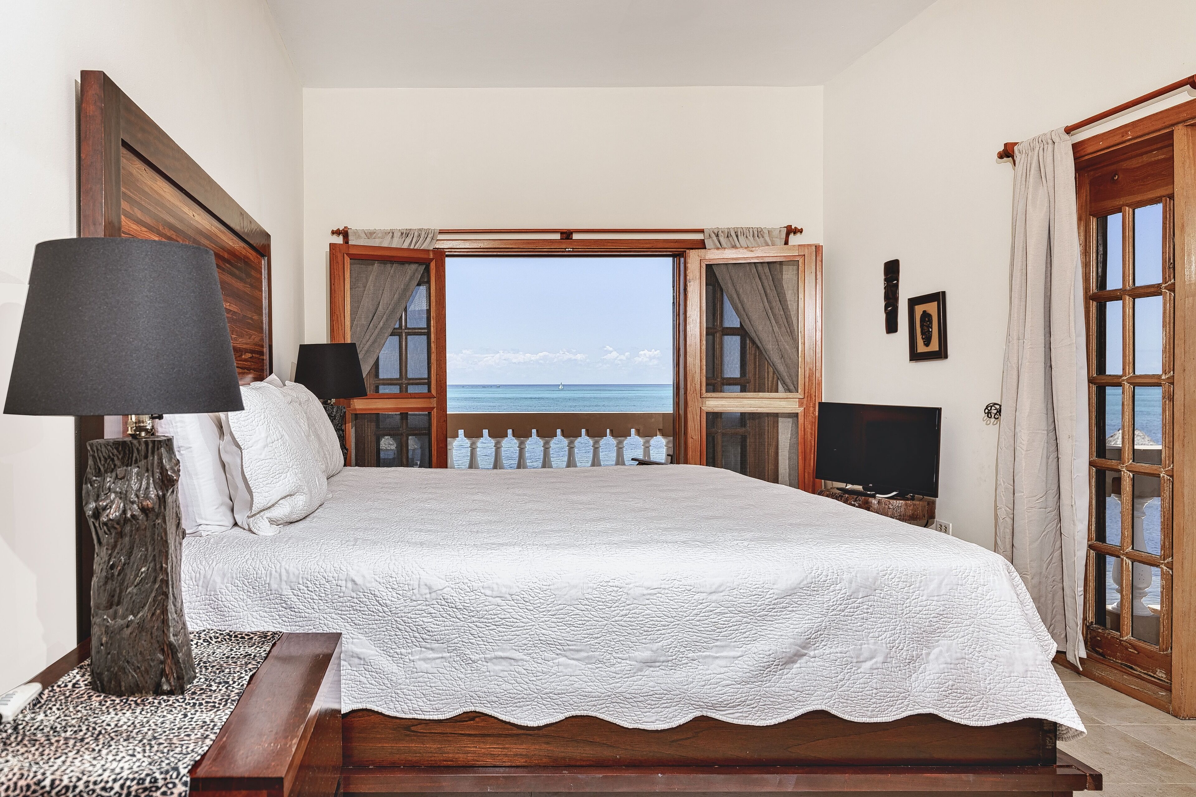 A bedroom with a large bed, a wooden headboard, a nightstand with a lamp, a TV, and a view of the sea through a window.