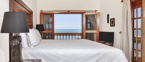 A bedroom with a large bed, a wooden headboard, a nightstand with a lamp, a TV, and a view of the sea through a window.