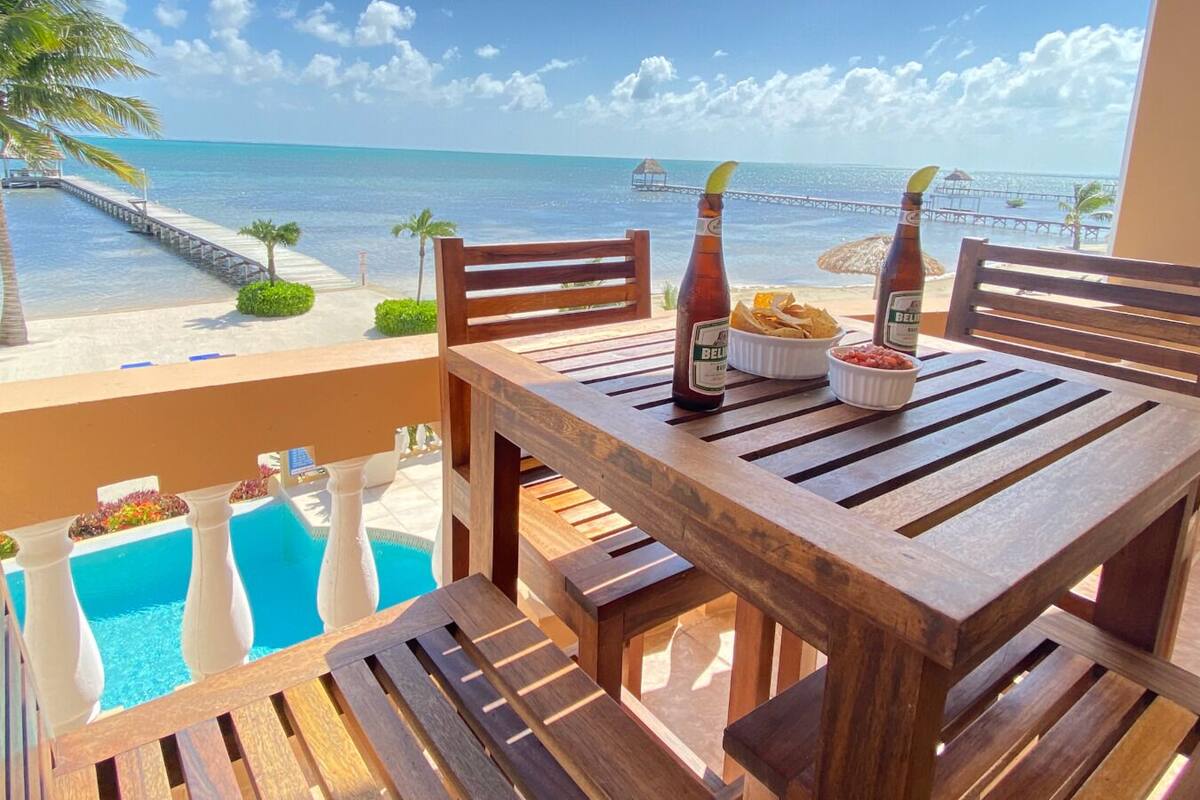 A wooden table with two bottles and a bowl of food on a balcony overlooking a pool and the ocean.
