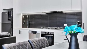 A modern kitchen with white cabinets, a black backsplash, stainless steel appliances, and a vase with blue flowers on the table. - Noir Cottage | Glenelg North | 3BR | Netflix| BBQ (Glenelg North)