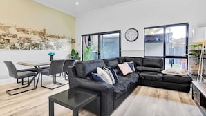 A modern living room with a black leather sofa, a dining table, and chairs. There is a large window with a view of the outside. - Noir Cottage | Glenelg North | 3BR | Netflix| BBQ (Glenelg North)