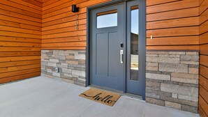Fachada de madera con puerta gris, detalles de piedra y una alfombrilla que dice 'hello'.