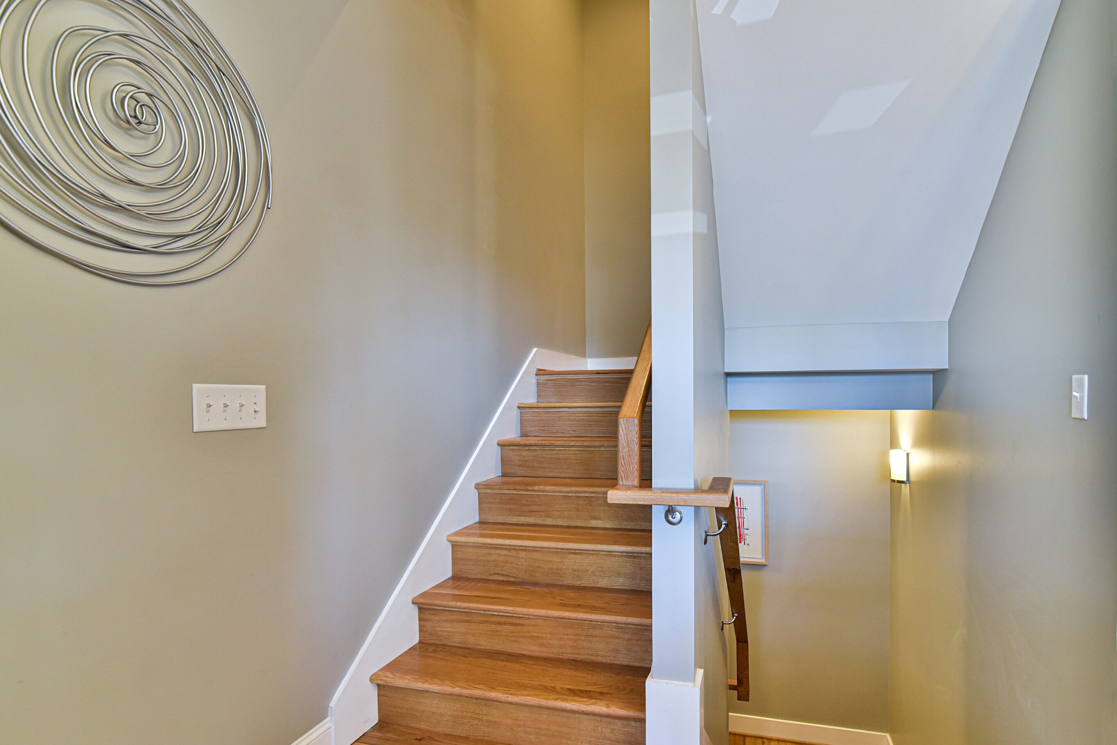 A wooden staircase with a handrail, a light switch, and a spiral metal decoration on the wall.