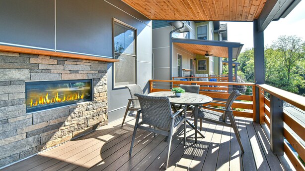 Una terraza de madera con un muro de piedra y una chimenea, mobiliario exterior y vistas a una casa.