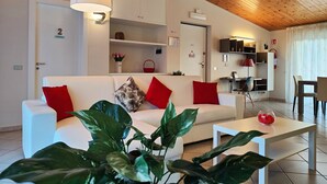 A modern living room with a white sofa, red cushions, a white coffee table, and large green plants. There is a wooden ceiling, a white door with a number, and a small kitchen area in the background.