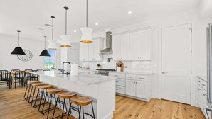 Una cocina moderna con armarios blancos, una isla grande con taburetes de bar, iluminación colgante y un reloj en la pared.
