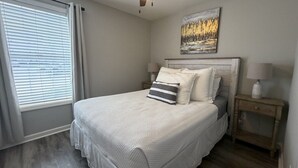 A bedroom with a bed, a nightstand, a lamp, a painting on the wall, and a window with blinds. - Walk to Toomer's! Ground-Floor 4 BR Auburn Condo (Auburn)