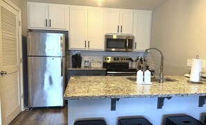 Una cocina compacta con armarios blancos, un refrigerador de acero inoxidable, un microondas y una encimera de granito.
