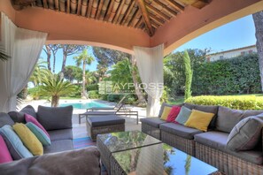 A covered outdoor seating area with wicker furniture, a glass-top table, and a view of a swimming pool and greenery. - Villa Pinede - heated pool (Grimaud)
