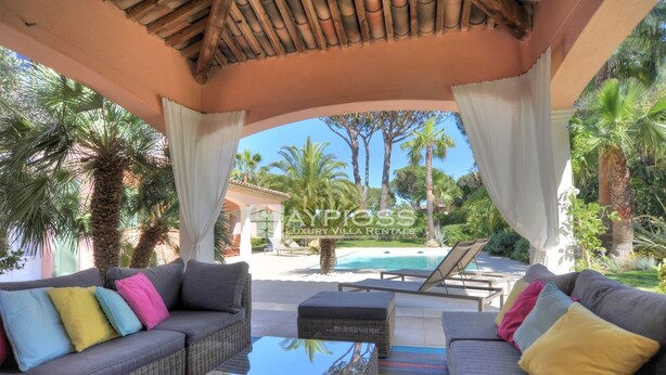 A covered outdoor seating area with wicker furniture, a glass table, and a view of a pool and palm trees.