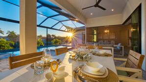 Un coin repas avec une table dressée pour le repas, un ventilateur de plafond et une vue sur une piscine et des palmiers.