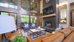 A spacious living room with leather sofas, a fireplace, and large windows offering a view of the outdoors. - New on Creek! HUGE Deck - Luxury Sleeps 15 - 4 Bedrooms (Broken Bow)