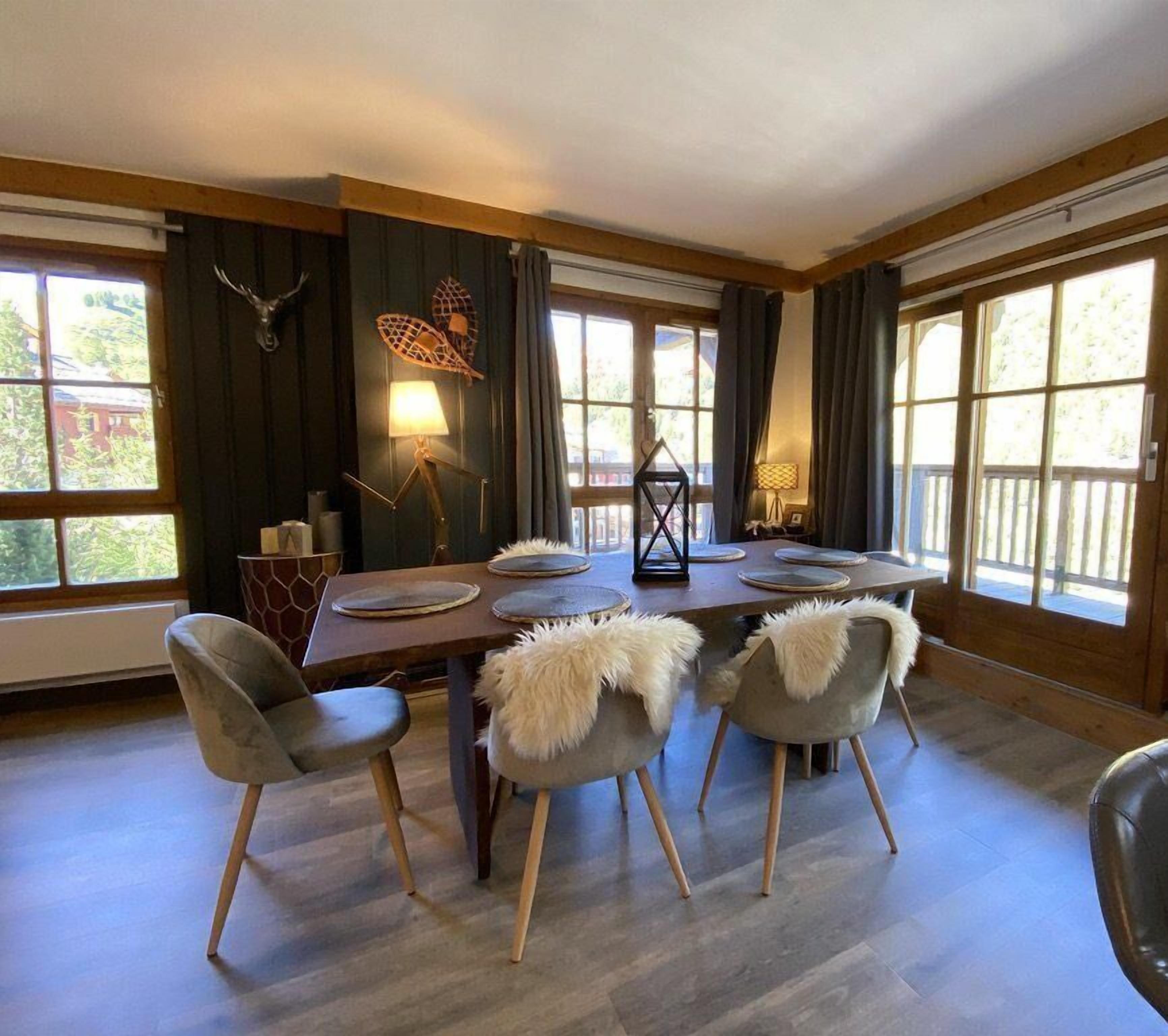 A dining room with a wooden table, grey chairs with fur throws, and large windows with curtains.