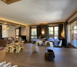 A spacious living area with a large wooden table, multiple seating options, and large windows allowing natural light.