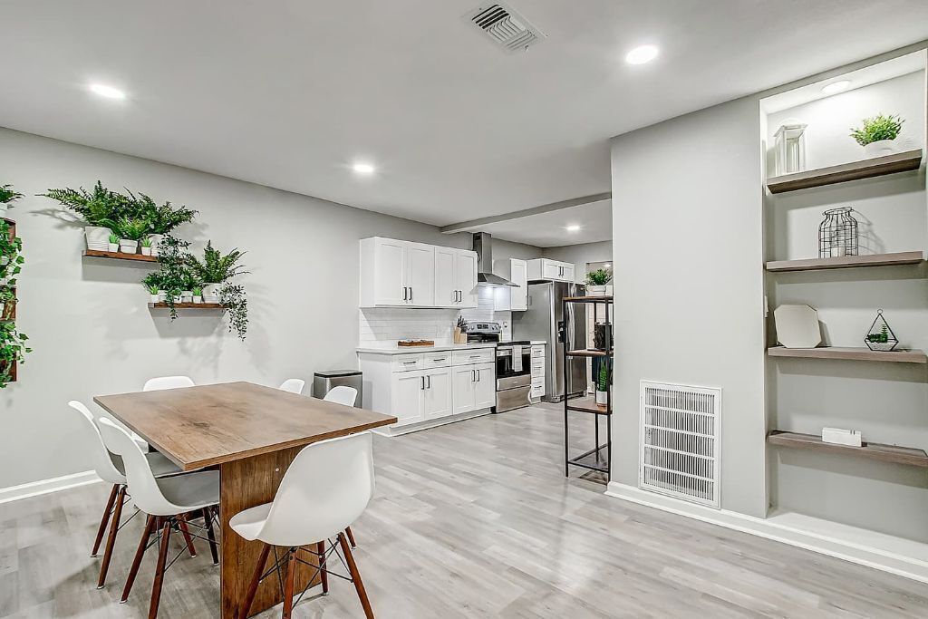 Una cocina moderna con armarios blancos, una mesa de comedor de madera y sillas blancas. Hay plantas en estantes y un radiador blanco en la pared.