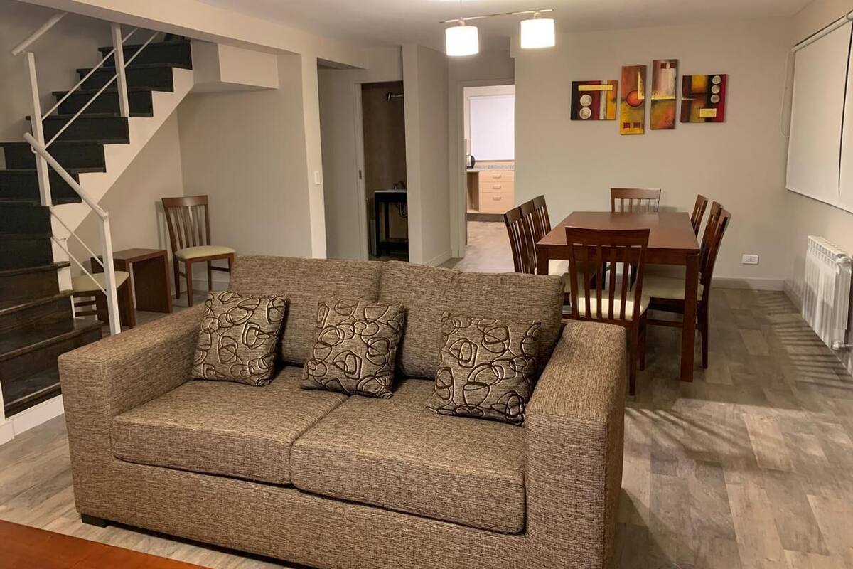 A living room with a sofa, a dining table, chairs, a staircase, and artwork on the wall.