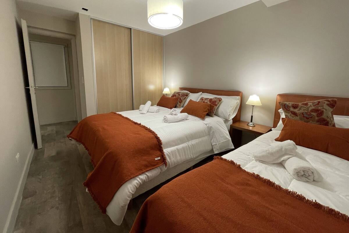 A hotel room with two beds, each with a brown blanket and white linens. There is a wooden headboard, a nightstand with a lamp, and a door leading to another room.