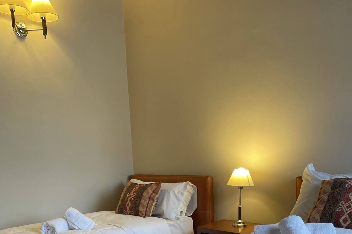 A hotel room with two beds, each with white linens and a patterned throw blanket. There are wall-mounted lamps providing soft lighting.