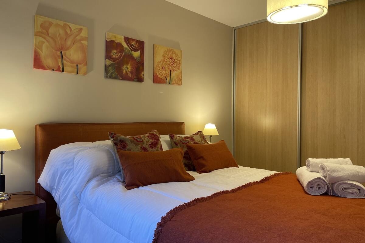 A neatly made bed with a brown headboard, white linens, and decorative pillows. There are three wall-mounted paintings above the bed, and a wooden wardrobe to the right.