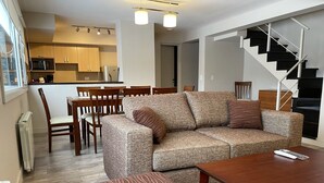 A modern living room with a sofa, ottoman, and coffee table, a dining area with chairs, and a kitchen in the background.