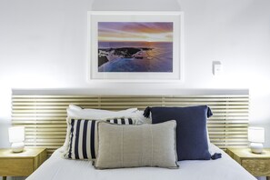 Een slaapkamer met een houten hoofdbord, een bed met veel kussens, een ingelijst foto van een zonsondergang en nachtkastjes met lampen.