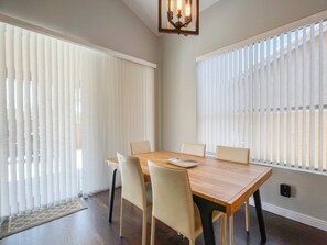 A dining room with a wooden table and six chairs, large windows with vertical blinds, and a modern chandelier.
