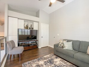 A living room with a grey sofa, a small grey armchair, a wooden entertainment unit with a flat-screen TV, and a patterned rug on the floor.