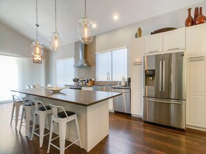 A modern kitchen with a central island, white bar stools, stainless steel appliances, and pendant lighting.