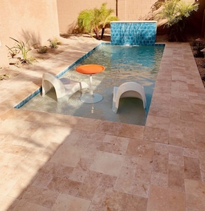 Una piscina con pared de azulejos azules, dos sillones blancos y una mesa naranja.
