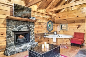 Interior de cabaña con chimenea de piedra, televisor montado en una viga de madera, lavamanos de piedra y una silla roja.