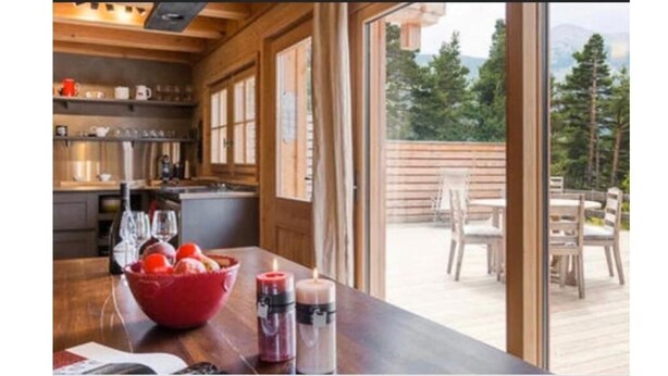 Interior de madera con cocina y comedor, ventanas amplias y vista a una terraza y árboles.