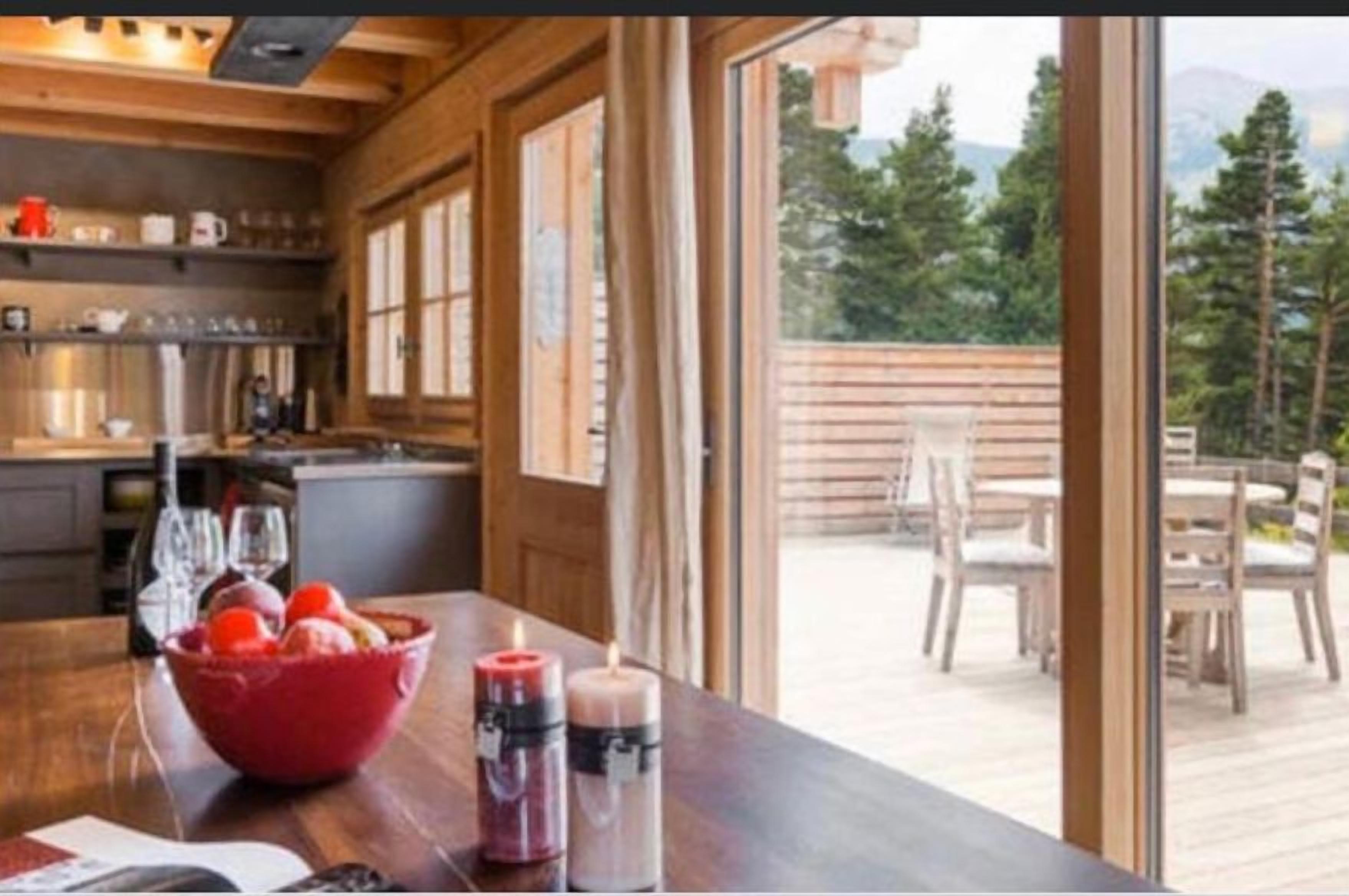 A wooden interior with a kitchen and dining area, large windows, and a view of a deck and trees.