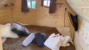 A small, cozy room with wooden walls, a bed with pillows and blankets, a window with curtains, and a mounted TV. - La Cabane Magique Des Fichettes (Sandillon)