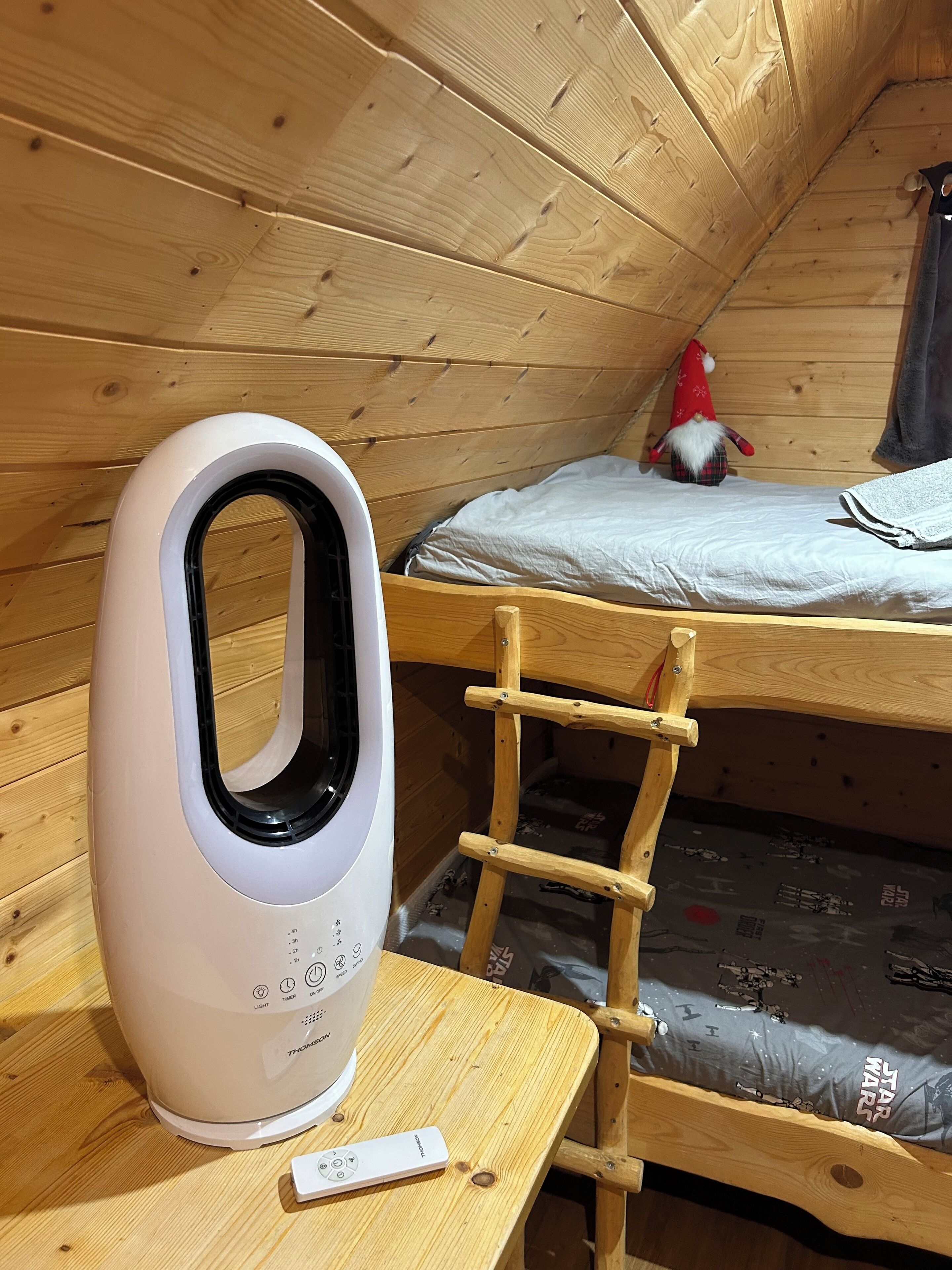 A wooden bunk bed with a ladder, a white electric heater, and a remote control on a wooden table.