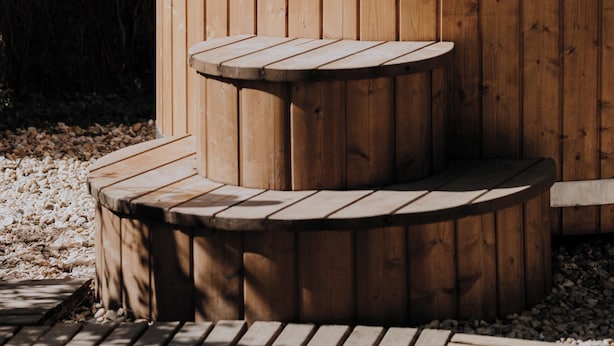 A wooden hot tub with two glasses of water, surrounded by a wooden deck and a hedge. - La Cabane Magique Des Fichettes (Sandillon)