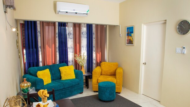 A living room with a blue sofa, a yellow armchair, and a teal ottoman. There is a dining table with plates and a vase. The room has a window with curtains, an air conditioning unit, and a door.