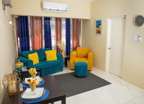 Una sala de estar con un sofá azul, un sillón amarillo y un reposapiés verde azulado. Hay una mesa de comedor con platos y un jarrón. La habitación tiene una ventana con cortinas, un equipo de aire acondicionado y una puerta.