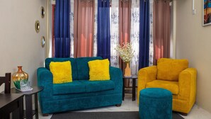 A living room with a teal sofa, a yellow armchair, and a teal ottoman. There is a vase with flowers on a side table.