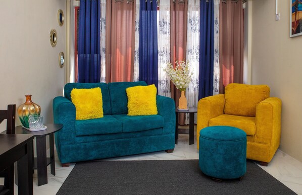 Una sala de estar con un sofá azul verdoso, un sillón amarillo y un reposapiés azul verdoso. Hay un jarrón con flores sobre una mesita auxiliar.